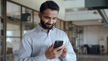 Happy indian ethnic businessman holding smartphone using mobile application program looking at cell phone working checking online digital data market corporate management tech standing in office.