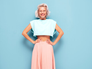 Portrait of young beautiful smiling female in trendy summer clothes. Sexy carefree woman posing near blue wall in studio. Positive blond model having fun and going crazy. Cheerful and happy