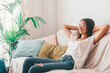 © Daniel - Happy young woman in eyeglasses relaxing on sofa with hands behind head in the living room of her house. Carefree and satisfied lady with torn jeans relaxing on couch at modern apartment.
