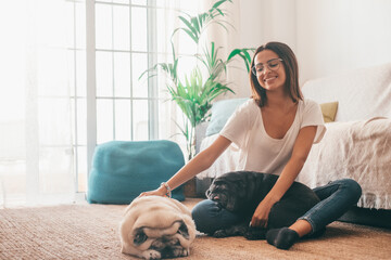 Portrait of beautiful woman in eyeglasses having fun with her pets pugs dogs sitting on floor in the living room of her house. Cheerful woman spending leisure time with her two cute pets dog at home.