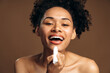 © speed300 - Portrait of charming multiracial curly woman with naked shoulders holding lip balm and preparing it putting into her lips. Isolated on brown background