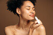 © speed300 - Close up cropped portrait of charming woman with naked shoulders holding lip balm. Isolated on brown background