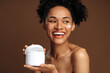 © speed300 - Attractive young woman holding jar with moisturizer to the camera while posing in studio over the brown wall. Stock photo