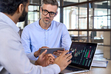 Diverse managers traders analysts discussing financial growth market at desk with laptop with graphs on screen using tablet device. Investors brokers analysing indexes online cryptocurrency stock.