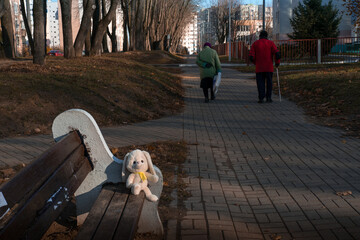 The poor elderly woman and the man walk along the path, they passed by the bench with the white toy hare that someone has left behind