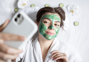 Wall Mural - Young woman with a cosmetic mask on her face makes a selfie on a smartphone.