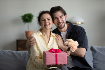 portrait of bonding young grownup son and sincere joyful mature retired mother sitting on couch, hol