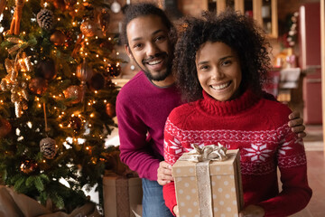 Wall Mural - Portrait of happy young African American couple pose with holiday presents near beautiful decorated Christmas tree at home. Smiling biracial man and woman exchange gifts celebrate New Year together.