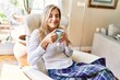 © Krakenimages.com - Young blonde woman drinking cup of coffee sitting on armchair at home.