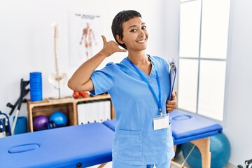 Wall Mural - Young hispanic woman with short hair working at pain recovery clinic smiling doing phone gesture with hand and fingers like talking on the telephone. communicating concepts.