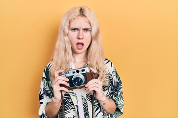 Sticker - Beautiful caucasian woman with blond hair holding vintage camera in shock face, looking skeptical and sarcastic, surprised with open mouth