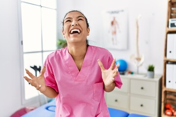 Poster - Young hispanic woman working at pain recovery clinic crazy and mad shouting and yelling with aggressive expression and arms raised. frustration concept.