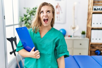 Poster - Young caucasian woman working at pain recovery clinic angry and mad screaming frustrated and furious, shouting with anger. rage and aggressive concept.