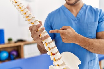 Sticker - Young hispanic man wearing physio therapist uniform pointing to anatomical model of vertebral column at clinic