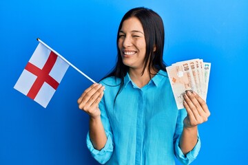 Poster - Young latin woman holding england flag and pounds banknotes winking looking at the camera with sexy expression, cheerful and happy face.