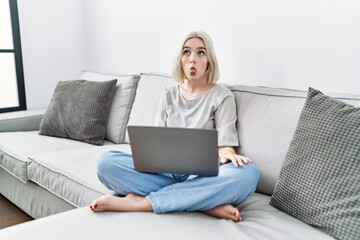 Sticker - Young caucasian woman using laptop at home sitting on the sofa making fish face with lips, crazy and comical gesture. funny expression.