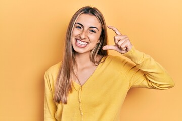 Canvas Print - Beautiful hispanic woman wearing casual yellow sweater smiling and confident gesturing with hand doing small size sign with fingers looking and the camera. measure concept.