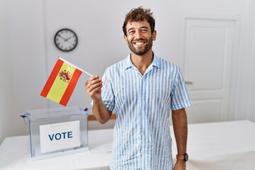 Sticker - Young handsome man at political campaign election holding spain flag looking positive and happy standing and smiling with a confident smile showing teeth