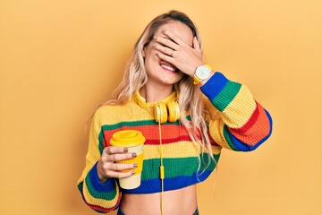 Sticker - Beautiful young blonde woman drinking cup of coffee wearing headphones smiling and laughing with hand on face covering eyes for surprise. blind concept.