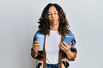 Wall Mural - Middle age hispanic woman drinking a cup of coffee and looking at the smartphone screen making fish face with mouth and squinting eyes, crazy and comical.