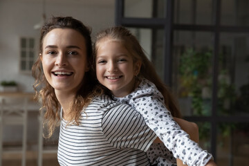 Wall Mural - Portrait of beautiful young mother giving piggyback ride to little child daughter, happy millennial mum and cute small kid girl having fun, entertaining together on weekend at home, looking at camera.