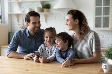 Wall Mural - Happy friendly young couple parents using cellphone with small kids, having fun entertaining online together, playing mobile games or watching funny photo video content in social network at home.