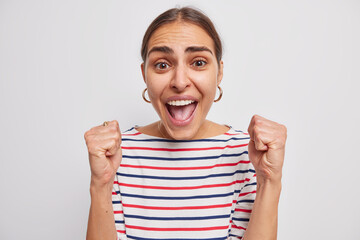 Wall Mural - Positive excited woman clenches fists and exclaims cheers over something shouts from joy boosts someone confidence encourages herself wears casual striped jumper isolated over white background.