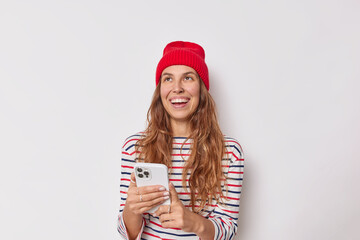 Wall Mural - Smiling beautiful young woman with happy expression uses mobile phone gadget wears red hat casual striped jumper isolated over white background. People modern technologies and lifestyle concept