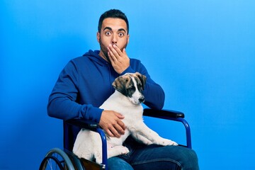 Poster - Handsome hispanic man with beard sitting on wheelchair with puppy dog covering mouth with hand, shocked and afraid for mistake. surprised expression