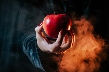 Woman as witch in black offers red apple as symbol of temptation, poison. Fairy tale, white snow wizard concept. Spooky halloween, cosplay. Smoke, haze background.