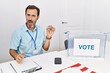 © Krakenimages.com - Middle age man with beard sitting by ballot holding i vote badge in shock face, looking skeptical and sarcastic, surprised with open mouth