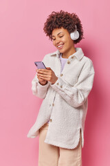 Wall Mural - Vertical shot of joyful woman with curly hair holds mobile phone chats online and listens favorite music in headphones wears stylish casual clothes isolated over pink background enjoys popular song