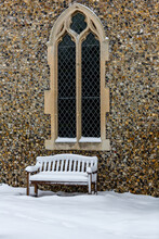 A Snow Covered Wooden Bench Outside Under A Large Lancet Church Window