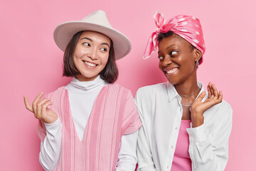 Wall Mural - Happy positive women have friendly relationships look gladfully at each other stands next to each other dressed in fashionable clothes isolated over pink background. Glad friends meet together