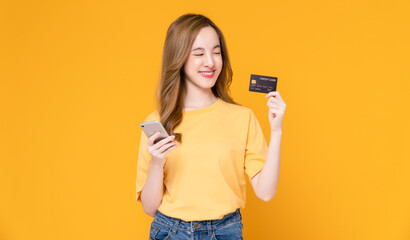 Wall Mural - Cheerful beautiful Asian woman holding smartphone and mockup credit card on orange background.