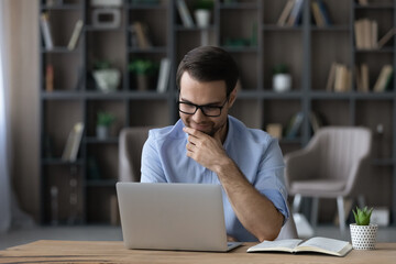 Poster - Smiling young businessman in eyewear looking at laptop screen, reading email with pleasant news, feeling satisfied with marketing research results, working on online project in modern office room.