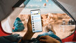 © Gorodenkoff - POV of a Tourist Vertically Holding a Smartphone and Checking Social Network Messages, Swiping a Finger Over Display. Traveller Resting in a Tent on Top of a Rocky Mountain and Flying Hot Air Balloons