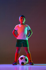 Wall Mural - Full-length portrait of boy, child, football player in uniform with ball posing isolated over gradient background in neon light.