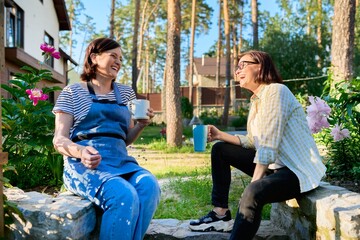 Two middle aged women sitting outdoors together in garden, backyard with mugs of tea