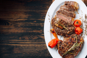 Wall Mural - juicy, well-done ribeye steaks on a white platter on a dark wooden table top with grammarine and fried cherry tomatoes and pepper