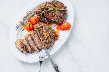 Wall Mural - juicy well-fried ribeye steaks on a white platter on a white marble surface with raspmarine and fried cherry tomatoes and peppers. steaks are sliced and ready to serve