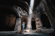 Woman tourist explores the mystical interior of the main hall of the Armenian Geghard Monastery. A ray of light falls on ancient bas-reliefs carved on the walls