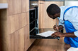 © alfa27 - Focused African Handyman Writing Information On Paper Document About Electric Oven Before Or After Repair, Side View On Confident Serious Guy Examining Inside Of Oven. Male In Blue Overalls, Workwear