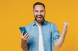 Young overjoyed cool happy caucasian man 20s wear blue shirt white t-shirt hold in hand use mobile cell phone chatting surfing do winner gesture isolated on plain yellow background studio portrait
