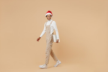 Wall Mural - Full body side view happy young african man wear Santa Claus red Christmas hat hold closed laptop pc computer isolated on plain pastel beige background studio. Happy New Year 2022 celebration concept.