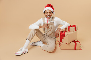 Full body young african man wear Santa Claus red Christmas hat sit near many present gifts boxes eat candy cane lollipop isolated on plain pastel beige background studio. Happy New Year 2022 concept.