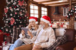 © insta_photos - Happy 50s older couple using digital tablet in living room on Christmas. Smiling mature grandparents wearing santa hats doing holiday online shopping having family video call relaxing on sofa at home.