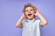Funny Child Boy Closing Ears Yelling By Amazement Isolated Over Purple Background In Studio. Cute European Caucasian Kid With Curly Hair In Casual Outfit T-shirt Looking Up, Screaming