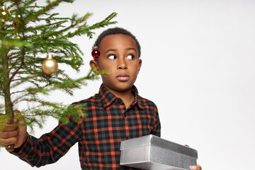 Wall Mural - Children, emotions and reactions. Cute lovely African boy in shirt making eyes bigger with shock, looking aside with worried face expression, holding decorated branch of pine and present box