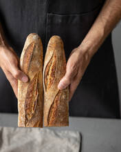An Adult European Male Baker Holds A Round Fresh Bread In His Hands. A Man In A Bakery Holds A Yeast-free Bread On Sourdough And A Baguette. Cool And Healthy Bread For The Whole Family. Vegan Food For